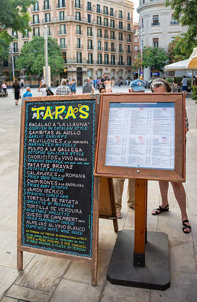 Barcelona, Spain - September  26, 2016 - A vibrant chalkboard menu showcases various tapas options, set against the backdrop of the Plaça Reial in Barcelona, Spain. People stroll by, enjoying the sunny day, while the historic buildings create a charming European ambiance. The enticing menu promises a delightful culinary experience on a summer day.