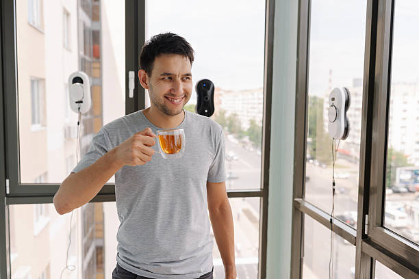 Young man enjoying cup of tea while robotic window cleaners work, showcasing convenience of modern smart home technology in contemporary apartment setting. Concept of cleanliness and housekeeping.
