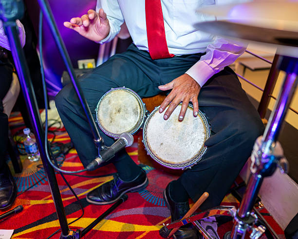 Over shoulder shot of Black male percussionist performing rhythmic beats on traditional conga drums while playing live music along with band in jazz club, illuminated by blue neon lights