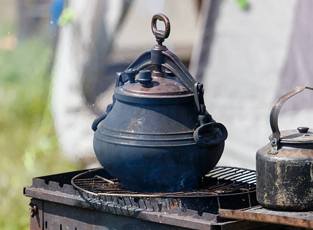 A large black metal pot containing white liquid hangs over an active wood fire inside a rustic, darkened structure. This captures the ancient process of cheese making or dairy heating in a mountain cabin