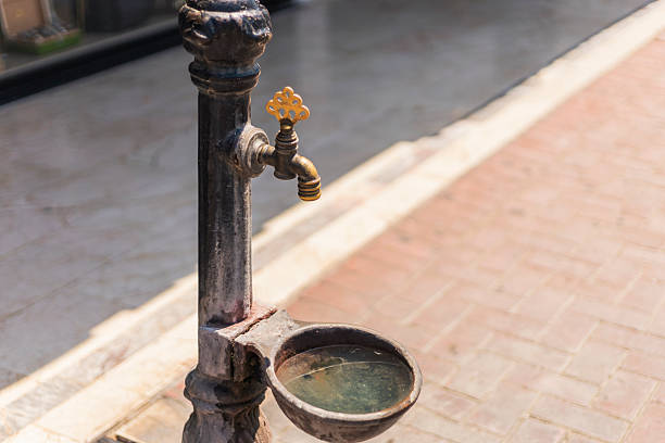 A vintage water fountain stands on a brick path, featuring a curved basin that holds clear water. The sun shines brightly, illuminating the surrounding urban landscape.