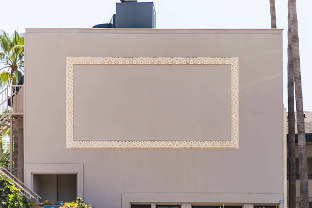 A beige building features an ornate wall frame design surrounding a blank area. Bright sunlight highlights the decorative elements, and palm trees add a tropical touch to the environment