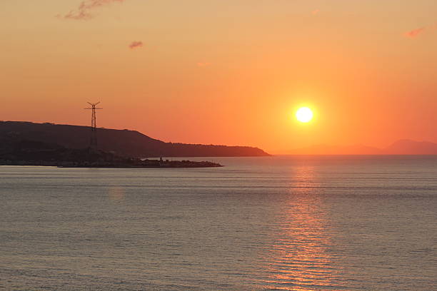 A breathtaking sunset over the ocean. The warm glow reflects on the calm water, with distant mountains and a tall transmission tower visible on the horizon.