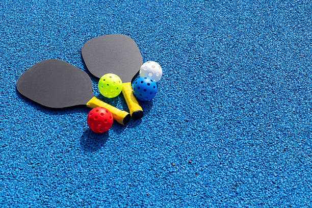 Medium shot of two pickleball rackets and four multicoloured balls laid on a blue sports court surface on a bright sunny day in Newcastle-upon-Tyne, North East England.