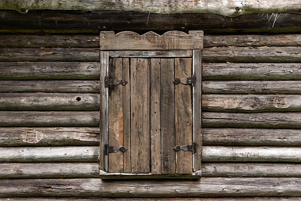 A vintage wooden window with closed shutter set against the textured exterior of a rustic log cabin, depicting simplicity and a timeless rural ambiance. A Northern Russian village architecture details