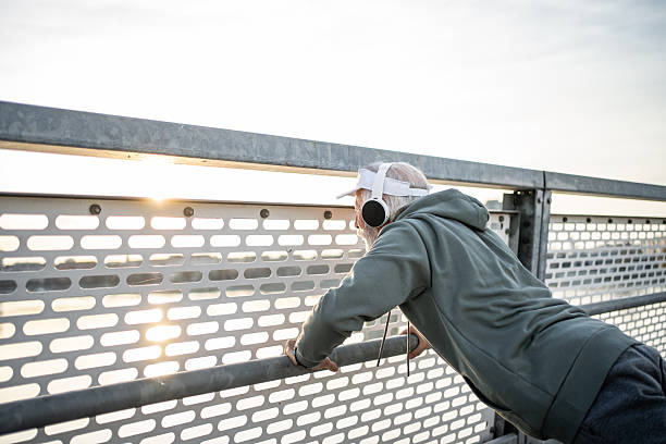 Senior man taking a break from exercising outdoors, resting against a bridge railing while enjoying the soothing sounds of music and the breathtaking sunset view