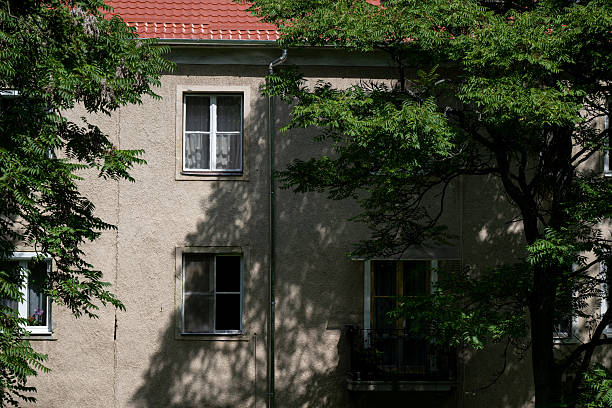 A residential building with a red tiled roof and satellite dish is partly framed by green trees. The window on the facade and the clear blue sky above create a calm and balanced suburban atmosphere.