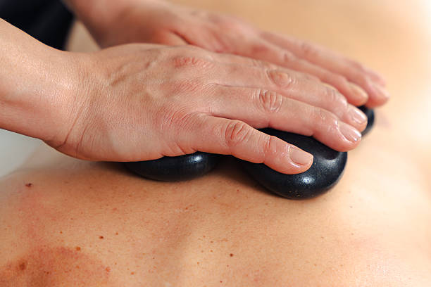 Professional massage therapist in green shirt placing hot stones on the bare back of an unrecognizable client during a relaxing spa treatment in a wellness center with a peaceful atmosphere .