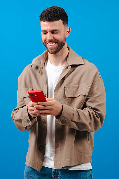 istock Bearded young man texting on red smartphone, wearing jacket on a blue background 2232342062