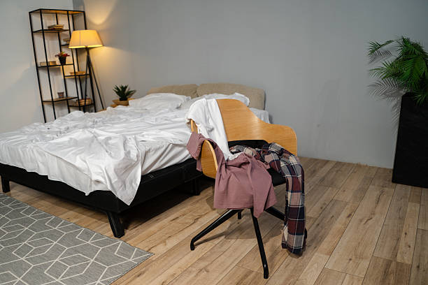 In a stylish bedroom filled with soft light, a bed stands unmade while clothes hang from a chair, reflecting a comfortable, carefree start to the day.