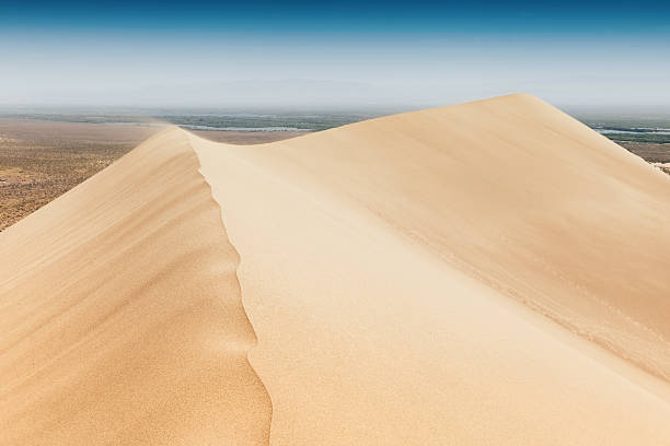 Strong wind blowing sand over the crest of a dune in the desert creating beautiful patterns on the sand, with vegetation and a river in the background