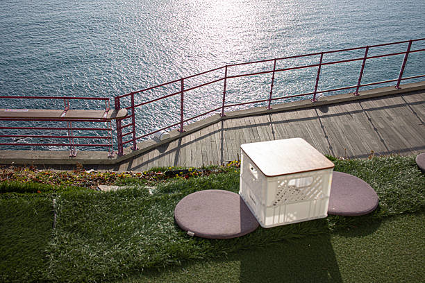 On a concrete terrace, a black wooden picnic bench faces the calm sea under a clear blue sky, sunny summer day. A metal railing frames the open view, emphasizing the peaceful and spacious atmosphere.