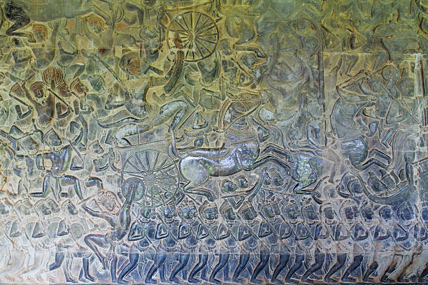 A Cambodian monk with yellow umbrella looking at bas-reliefs at the outer walls of the Bayon, Angkor Thom, Cambodia. The bas-reliefs show scenes from daily life back in the 12th century. At the top section, monkeys are visible in the tree-tops, while the lower panels show various types of cooking, with grilling of fish in braziers. Monks are not supposed to use sunglasses, which is where an umbrella is useful.
