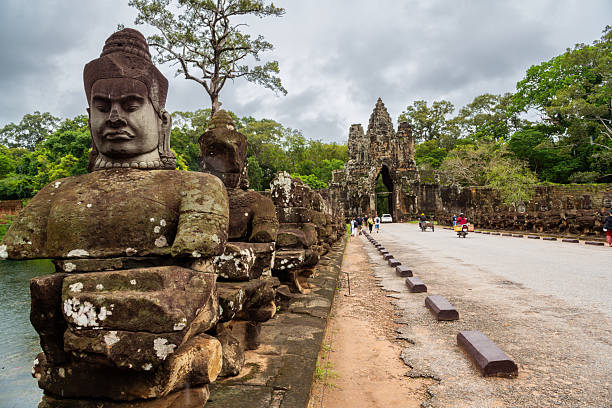 A serene view of the historical South Gate of Angkor Thom in Cambodia, showcasing ancient statues and intricate architectural details surrounded by lush greenery under a cloudy sky.