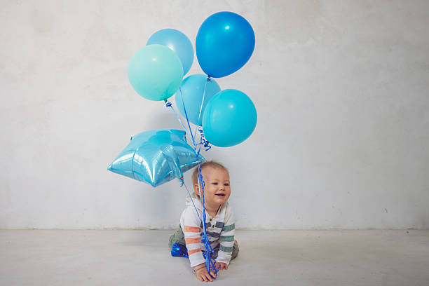 A playful photo of a baby holding onto colorful balloons, set against a simple white cement wall. A bright and joyful moment full of movement and charm.