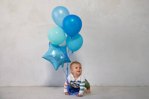 A playful photo of a baby holding onto colorful balloons, set against a simple white cement wall. A bright and joyful moment full of movement and charm.