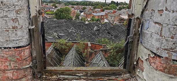 Urban view from broken window overlooking houses. Rotting frame missing glass, crumbling brickwork opening framing town below. Derelict wooden surround with collapsing roofs and apocalyptic overgrowth covering city beneath. Cityscape through open window, townscape falling apart, abandoned looking like UK slums. Looking through window down on ruins of industrial mill factory.