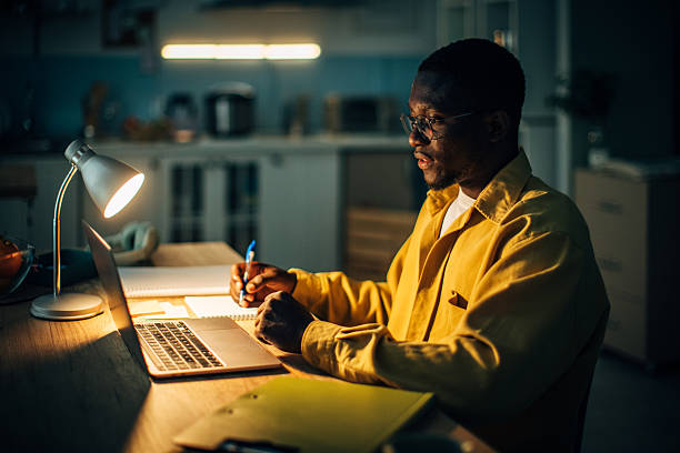 istock Young black man working on a laptop at home during the night with focused concentration and a warm desk lamp illuminating the space 2232373646