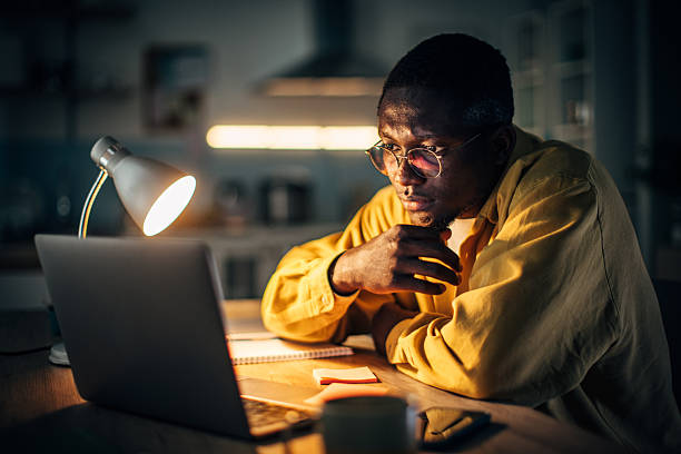 istock Young black man studying at home during nighttime with focused expression and warm desk lamp lighting 2232373729