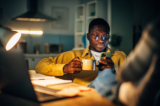A young black man is sitting comfortably at home in the evening. He holds a cup in one hand while focused on his smartphone, surrounded by warm light.