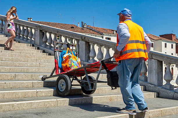 1 july 2025 Venice Italy: a porter in Venice, Italy, wearing a high-visibility orange vest and blue cap, pushing a two-wheeled cart loaded with colorful bags up the stone steps of a bridge. In the background, a young woman descends the stairs while talking on the phone. The scene illustrates the unique logistics of Venice, where goods are transported manually due to the absence of cars, showcasing the daily effort of local workers under the summer sun.