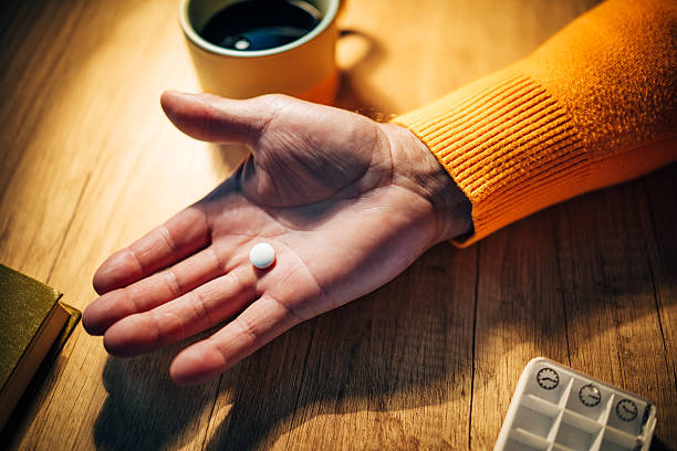 A senior man sits at a table in his cozy home, holding a pill in one hand while a cup of coffee sits nearby. The setting is calm and intimate, reflecting a quiet night.