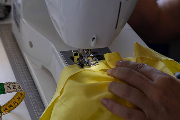 Hands work diligently on a sewing machine, creating a new piece with vibrant yellow fabric. A tape measure lies nearby, emphasizing the precision of the craft.