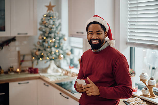 istock Happy black man wearing Santa cap while using cell phone at home and looking at camera. 2232378961