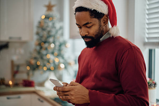 istock Black man wearing Santa cap while text messaging on Christmas at home. 2232378979