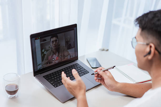 istock Asian man in glasses and earbuds gesturing hands while speaking on laptop video call at bright home office desk. Suitable for remote meetings, webinars, and online collaboration lifestyle themes 2232380836