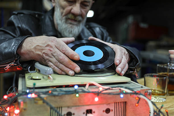 Elderly Man with Beard and Black Beret Holding Vinyl Record and Placing It on Turntable Surrounded by Festive Lights. Concept of Retro Music, Vintage Lifestyle, and Nostalgia for Analog Sound.