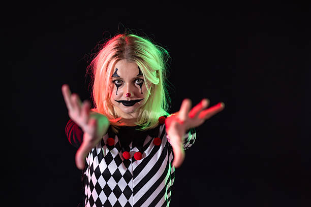 Woman dressed as a clown/harlequin, standing in front of a black studio background. She is looking at the camera with a creepy expression, reaching her hands out towards the camera.Videos are available similar to this scenario.