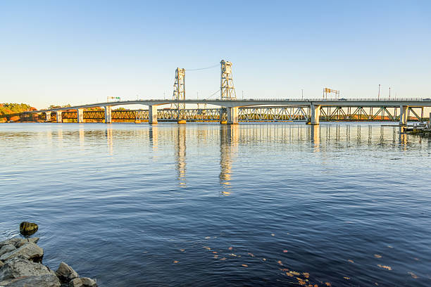 Highway bridge running parallel to a railway lift bridge over a mighty river at sunset in autumn. Reflection in water. Bath, ME, USA.