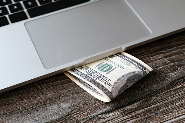 Stack of 100 dollar bills lying under laptop. Conceptual image of digital money. Selective focus.