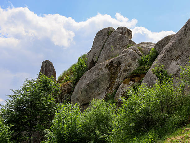 istock Massive stone boulders form majestic cliffs covered in moss and lush green vegetation. Below them are green bushes and trees, with a blue sky and white clouds shining above. 2232389793