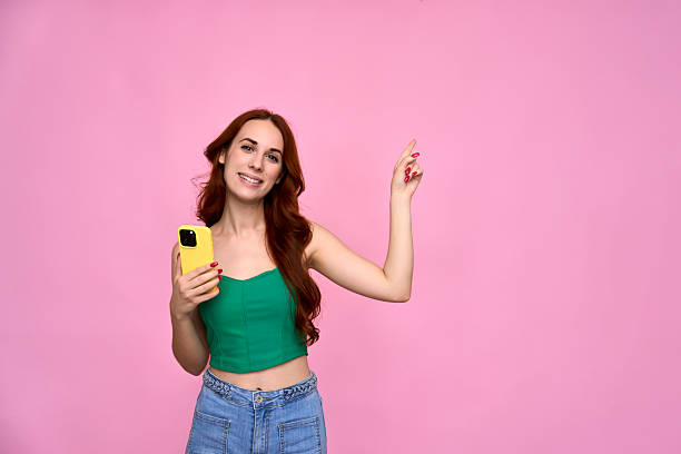 istock Young woman pointing up while holding smartphone on pink background 2232397668