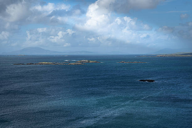 Overlooking the ocean. The mountains of Mayo County are in the background. The sea with small rocky islet is rippled by a strong wind. The sky is scattered with clouds.