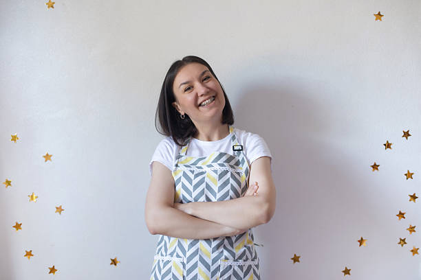 Smiling young woman with braces wearing a patterned apron stands against a white wall decorated with golden stars, arms crossed, radiating joy and confidence indoors.
