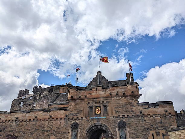 Edinburgh, UK - July 18 2025: The Royal Standard of Scotland and other flags fly above the historic stone gatehouse entrance of Edinburgh Castle against a partly cloudy blue sky