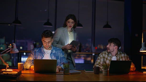 istock Two managers typing laptops at night desk. Lady entering office assisting team 2232417003