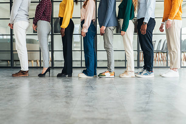 This photo shows a diverse group of people standing in a straight line, side by side, facing to the right. Only the lower halves of their bodies are visible, highlighting a variety of professional attire and footwear. The setting appears to be a modern office with a glass wall in the background, suggesting a business or corporate environment. Business people waiting in line to enter on a business conference.