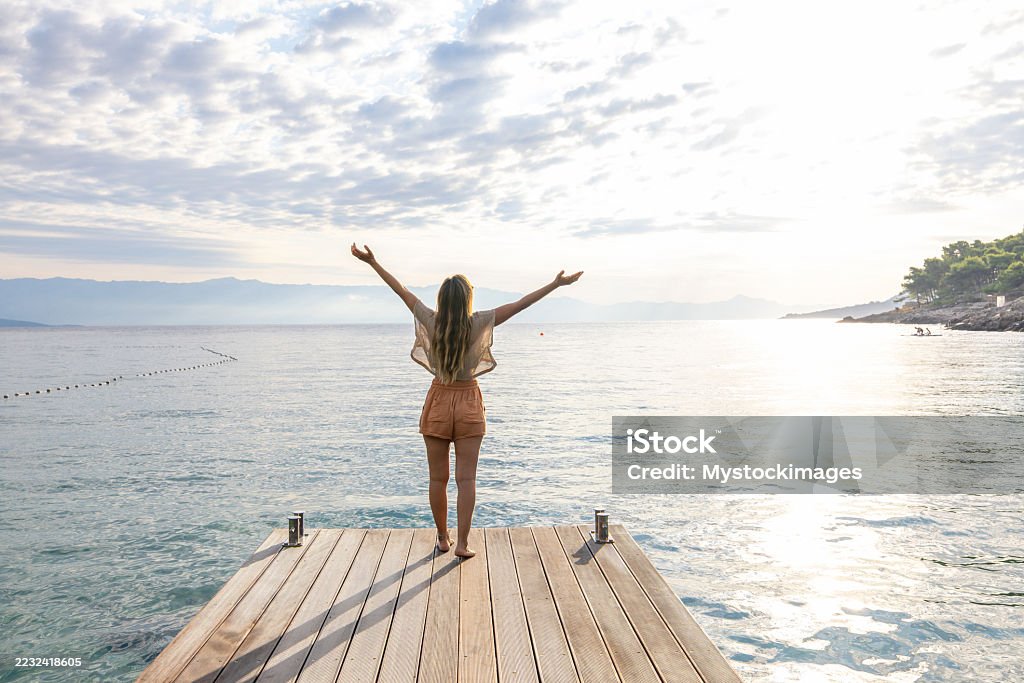Woman Standing on Dock with Raised Arms Young woman with long hair standing barefoot on wooden dock facing the sea with arms raised. Person enjoying nature and freedom by the water. Outdoor relaxation and travel concept. 35-39 Years Stock Photo Woman Standing on Dock with Raised Arms Young woman with long hair standing barefoot on wooden dock facing the sea with arms raised. Person enjoying nature and freedom by the water. Outdoor relaxation and travel concept. 35-39 Years Stock Photo
