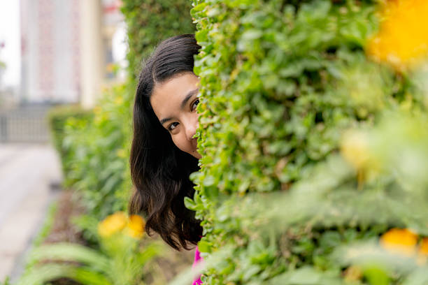Young woman peeking playfully from behind a lush hedge, enjoying a moment of relaxation and fun in a vibrant garden setting