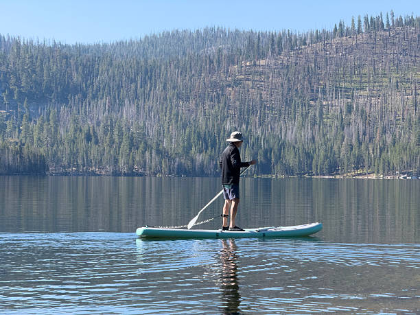 Man with hat, longsleeve black shirt and shorts standing up on paddle board on calm lake in the mountains. California Sierra Mountain Range