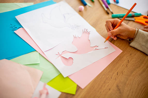 Kid drawing a stork carrying a baby on a white sheet placed on pink and colored sheets, using an orange pencil