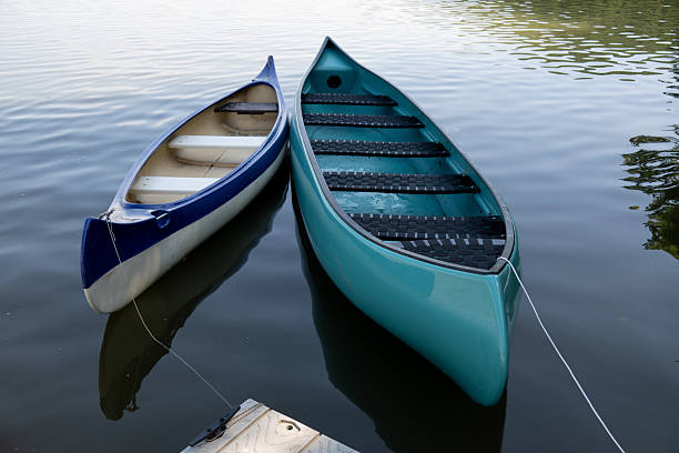 Two colorful canoes, one blue and one turquoise, are tied to a dock and gently floating on calm water. Their reflections create a serene and peaceful atmosphere, evoking a sense of leisure and outdoor adventure. The scene highlights relaxation, boating, and connection with nature.