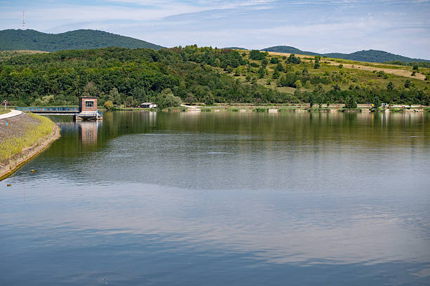A peaceful lake reflects the surrounding green hills and blue sky on a clear summer day. A small wooden dock structure extends into the water, adding a rustic element to the serene landscape. The scene conveys tranquility, nature, and relaxation in a rural environment.