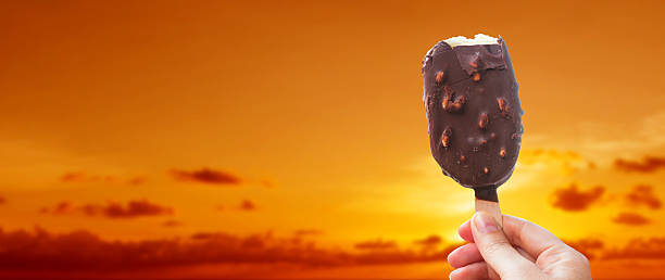 istock Chocolate covered ice cream bar with silhouetted sunset summer at the beach, during the holidays. Hand holds delicious chocolate and nuts almond ice cream bar on a stick. 2232436699