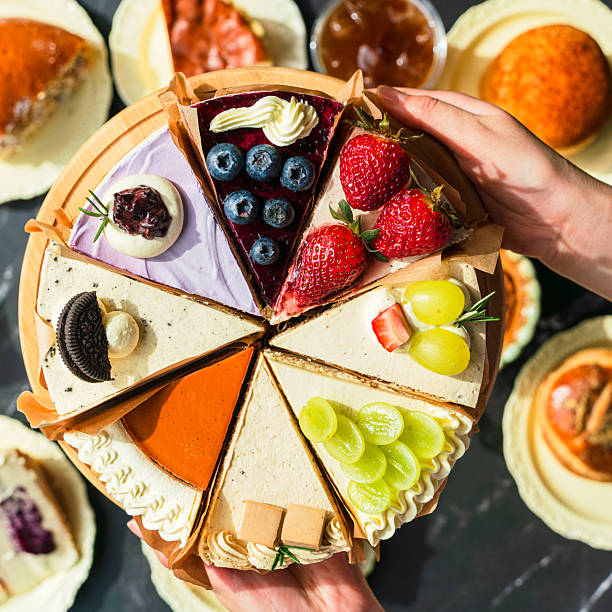 A wooden platter with assorted colorful cheesecake slices, surrounded by various desserts, held by a human hand against a dark background.