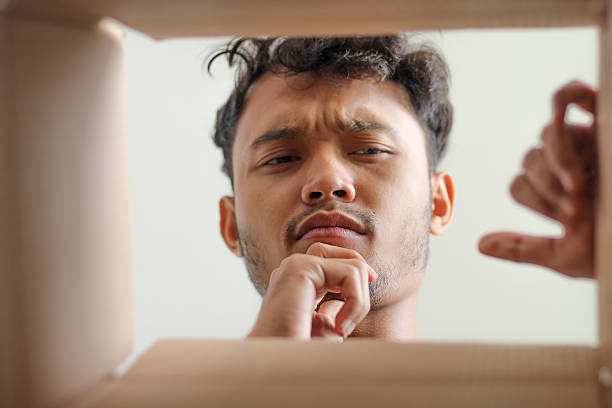 istock Young Asian Man Looking Into Box With Thoughtful Or Confused Expression Hands Reaching 2232443567
